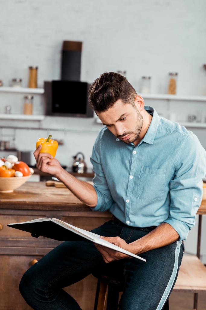 επικεντρώθηκε νεαρός άνδρας κρατά φρέσκο πιπέρι και διαβάζοντας το βιβλίο μαγειρικής στην κουζίνα - Φωτογραφία, εικόνα