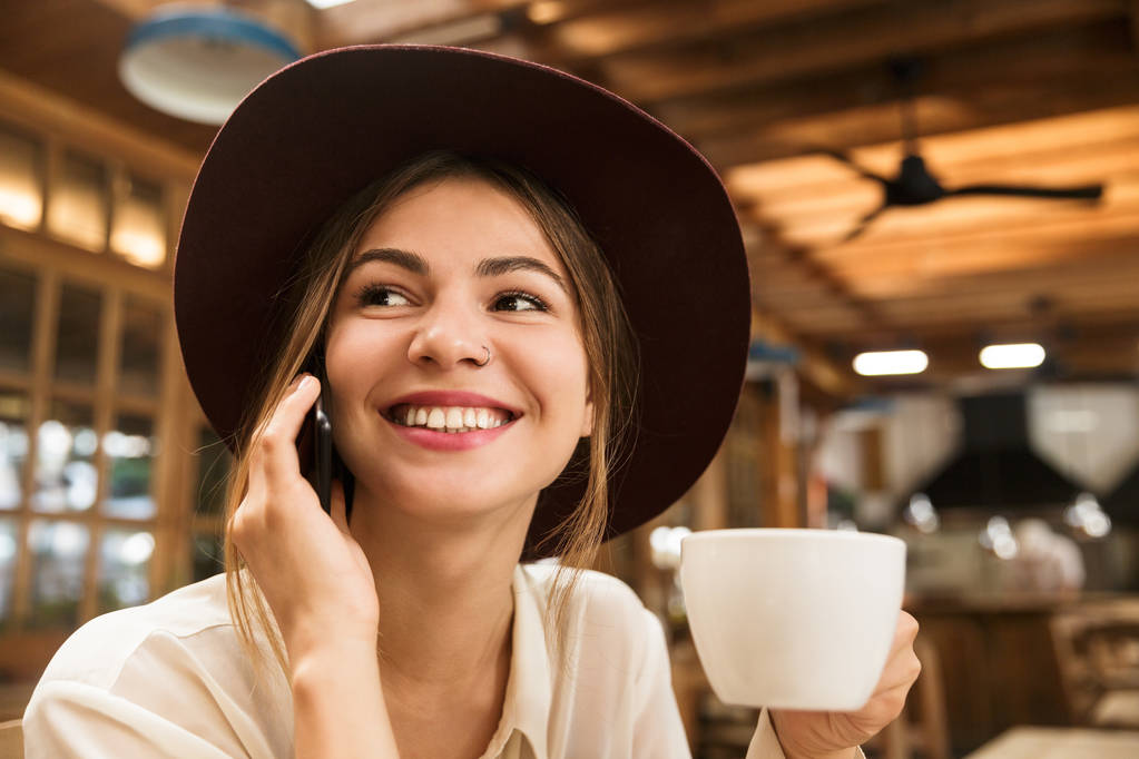 Nahaufnahme eines glücklichen hübschen Mädchens mit Hut, das drinnen am Cafétisch sitzt, eine Tasse Tee in der Hand hält und mit dem Handy telefoniert - Foto, Bild