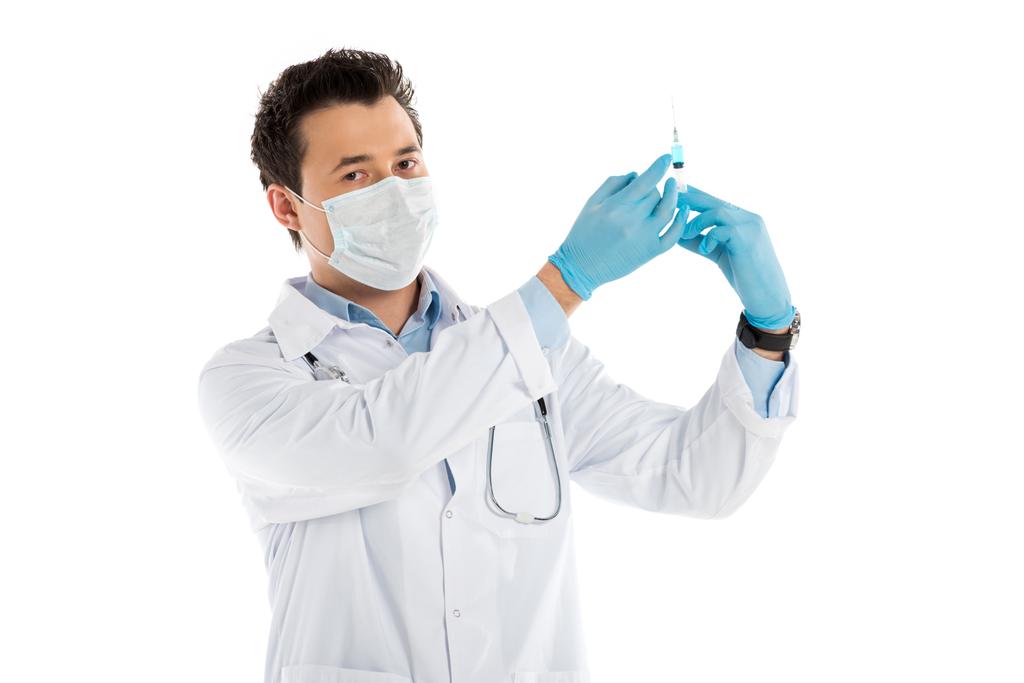 focused male doctor in rubber gloves and mask holding syringe and looking at camera isolated on white - Photo, Image