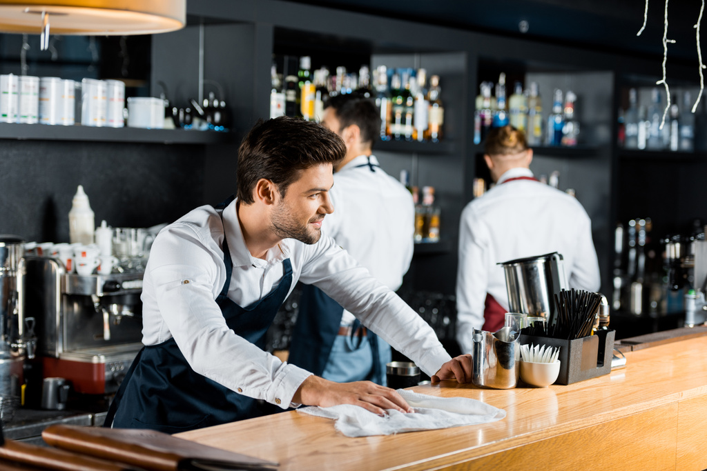 adult smiling barman cleaning wooden counter with cloth  - Photo, Image