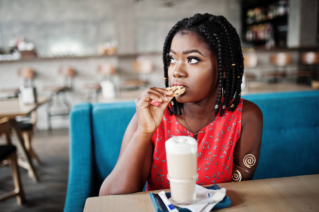 Atractiva mujer afroamericana sentada en la mesa en la cafetería con café con leche y comer galletas
. - Foto, imagen