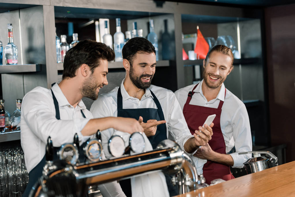 guapo adulto barmen sonriendo y usando smartphone en el lugar de trabajo
  - Foto, imagen