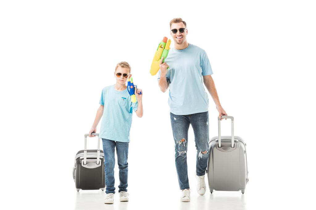 Isä ja poika kävelevät aurinkolaseissa ja pitävät vesipyssyjä ja matkatavaroita eristyksissä valkoisella
 - Valokuva, kuva