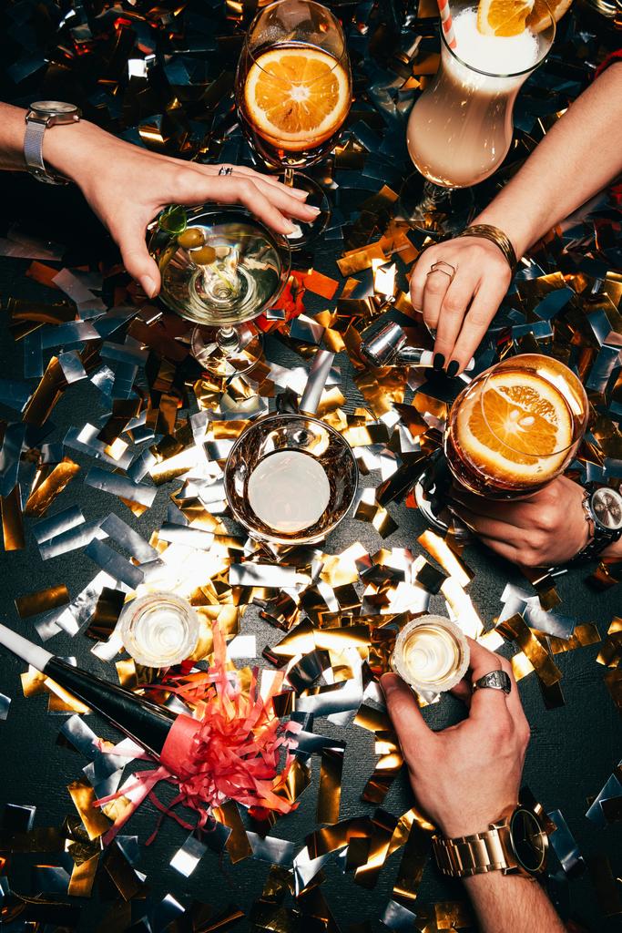 abgeschnittenes Bild von Freunden mit Luxusuhren, die Alkohol am mit goldenem Konfetti bedeckten Tisch feiern - Foto, Bild