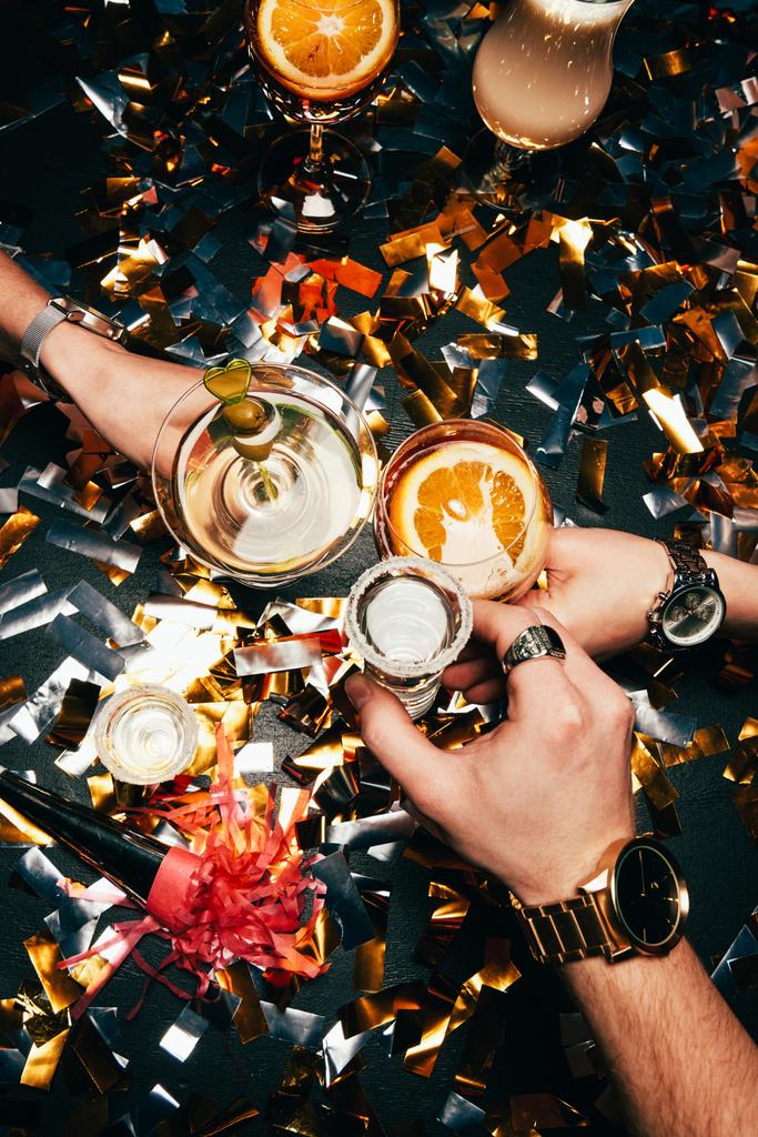 Μερική άποψη των φίλων με πολυτελή ρολόγια τσούγκριζαν από γυαλιά με οινόπνευμα πάνω από το τραπέζι καλυμμένο με χρυσές κομφετί - Φωτογραφία, εικόνα