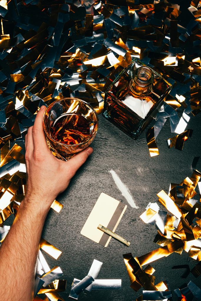 visão parcial do homem com copo de uísque perto da cocaína com notas e cartões de crédito enrolados na mesa coberta por confetes dourados
 - Foto, Imagem
