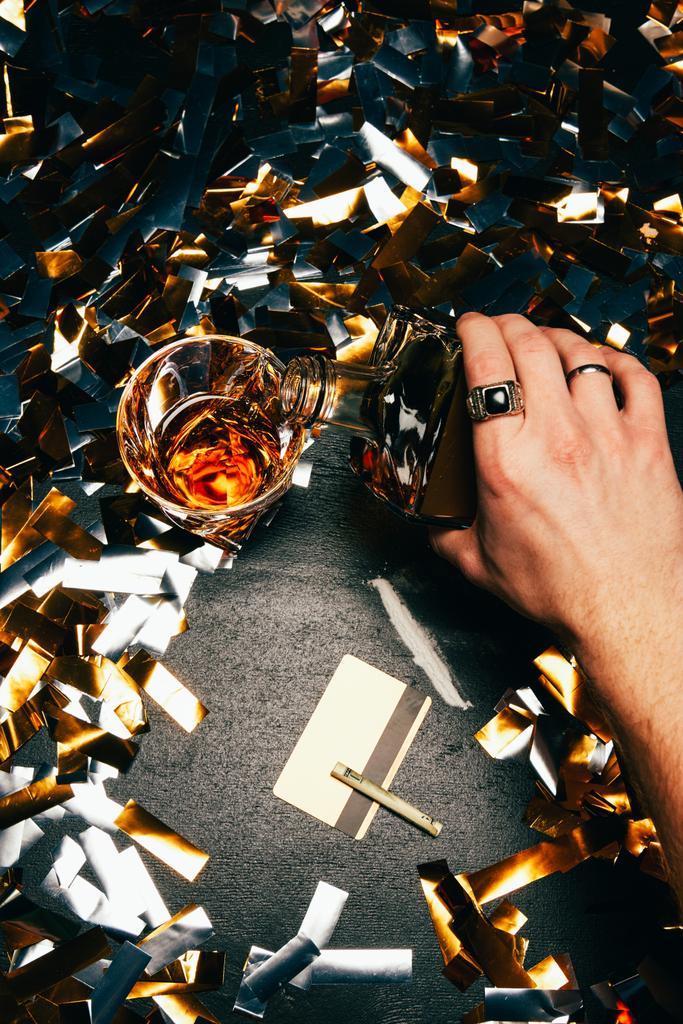 skadrowana człowiek wylewanie whisky do kieliszka w pobliżu kokainy z walcowanych banknotów oraz kartę kredytową na stół przykryty przez złotym konfetti - Zdjęcie, obraz