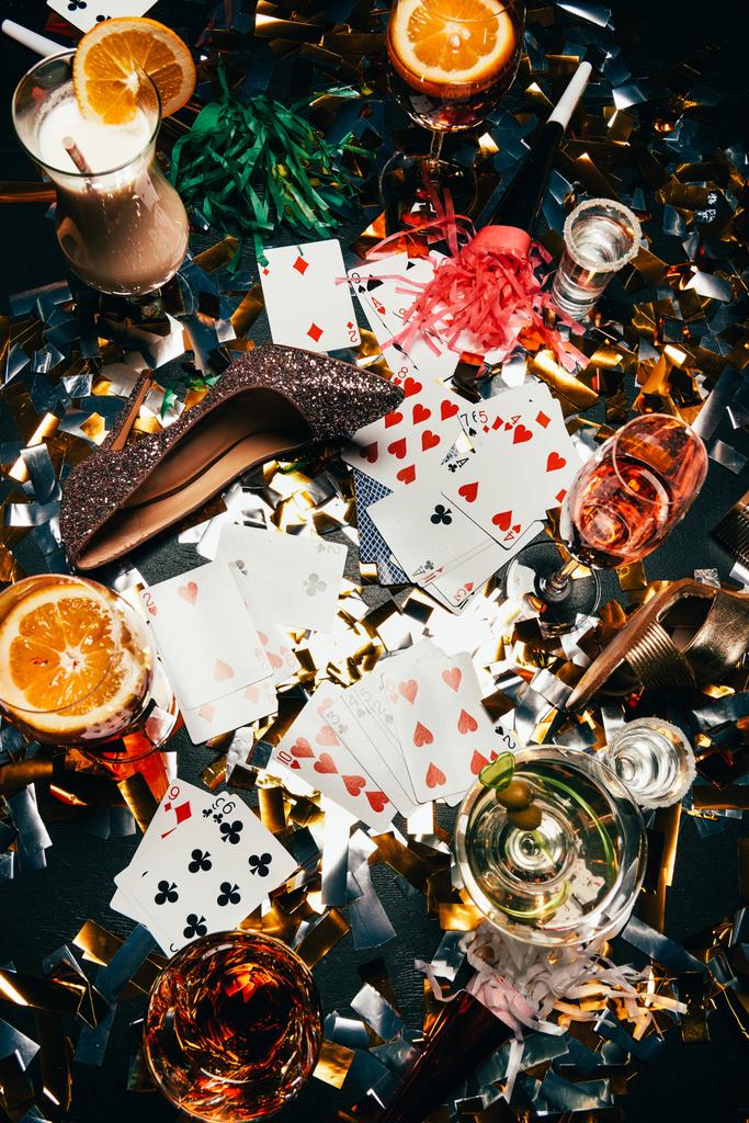 top view of female shoes on high heels, playing cards, alcoholic cocktails and party horns on table covered by golden confetti  - Photo, Image