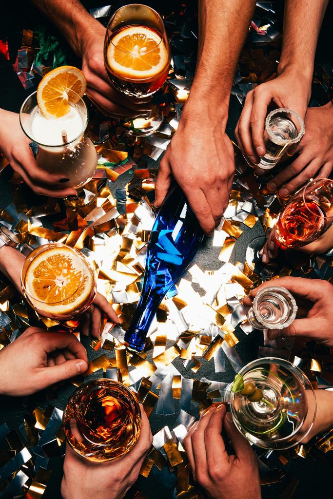 частичный взгляд друзей с различными алкогольными коктейлями, играющих в бутылку за столом, покрытым золотыми конфетти
  - Фото, изображение