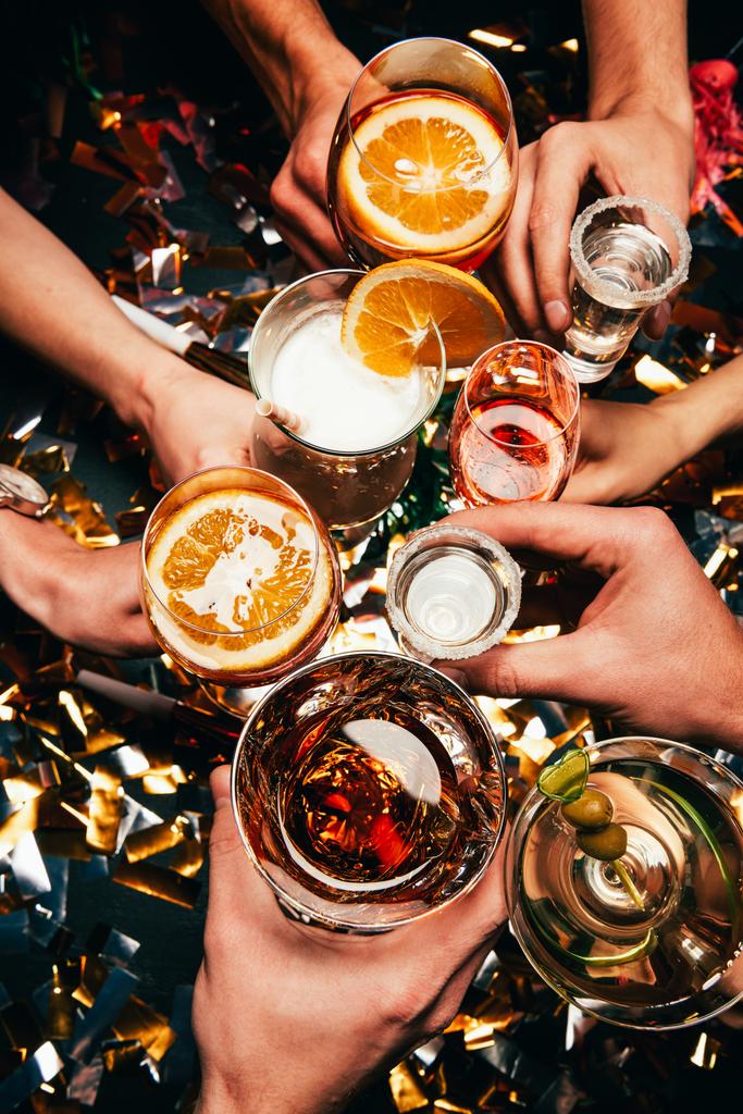 bijgesneden afbeelding van vrienden rammelende door bril met verschillende alcoholische cocktails aan tafel gedekt door gouden confetti  - Foto, afbeelding