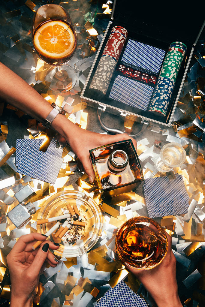 abgeschnittenes Bild von Frauen, die mit Alkohol, Zigaretten und Pokerchips feiern, mit Spielkarten auf einem Tisch, der von goldenem Konfetti bedeckt ist  - Foto, Bild