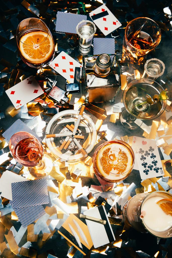 top view of cigarettes, alcoholic cocktails, playing cards, rolled banknote, credit cards and cocaine on table covered by golden confetti  - Photo, Image