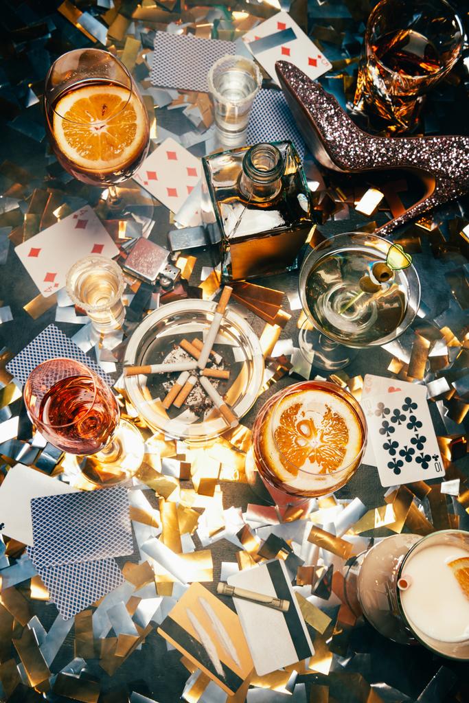 zvýšená pohled alkoholické koktejly, hrací karty, cigarety, válcované bankovky, kreditní karty a kokainu na tabulka překrytá zlaté konfety  - Fotografie, Obrázek