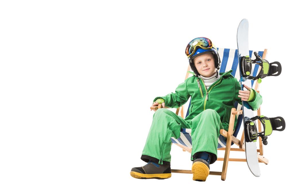 Frühchen Junge im grünen Skianzug mit Snowboard sitzt im Liegestuhl isoliert auf weiß - Foto, Bild