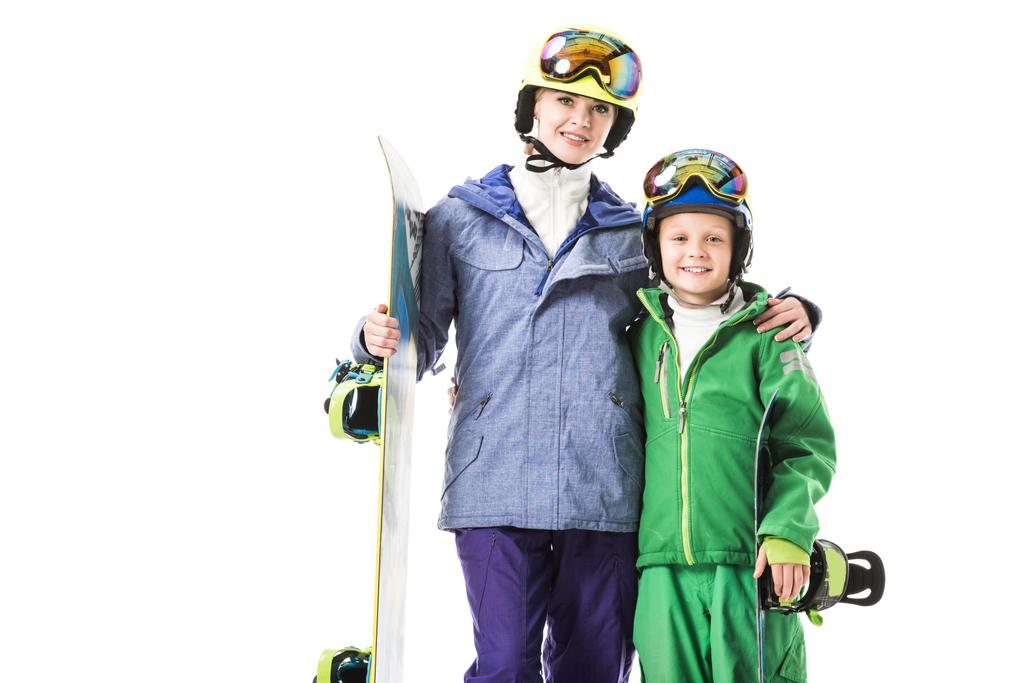 Lächelnde Mutter und frühpubertärer Sohn in Skianzügen mit Snowboards, die sich umarmen und isoliert auf weiß in die Kamera schauen - Foto, Bild