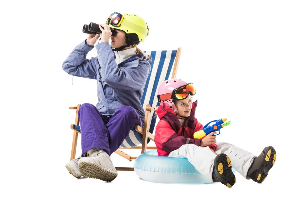 Kobieta w kombinezon narciarski z lornetki siedząc w leżaku, podczas gdy córka siedzi na pierścień pływania na białym tle pistoletem na wodę - Zdjęcie, obraz