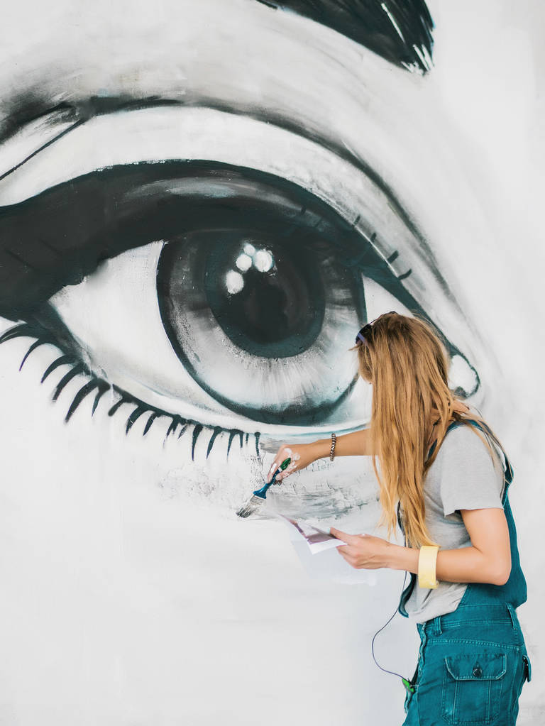 美しい少女は、都市の通りの壁にスプレーで大きな女性の顔の落書きを作るします。創造的な芸術。絵を描くデニム オーバー オールで才能のある学生 - 写真・画像