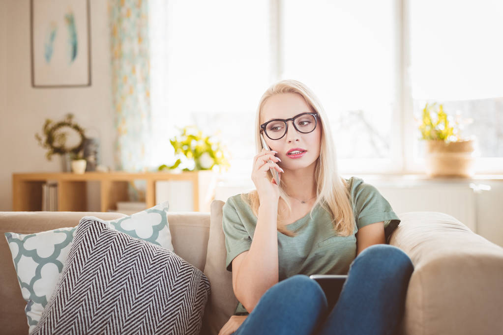 Mujer joven preocupada sentada en el sofá en casa y hablando por teléfono móvil
.  - Foto, Imagen