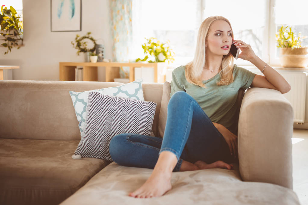 Huolestunut blondi nuori nainen istuu sohvalla olohuoneessaan ja puhuu matkapuhelimella
.  - Valokuva, kuva