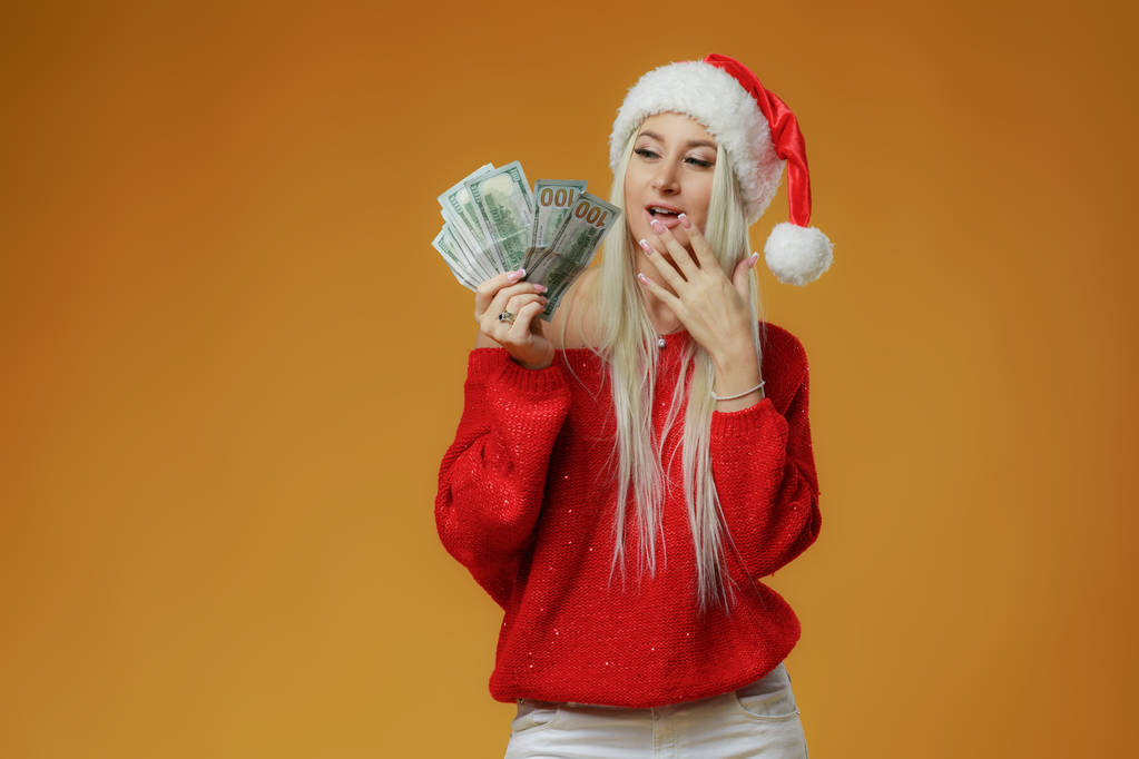 Pretty caucasian young happy woman in red Santa hat holding gift box and money banknotes on yellow background. Won cash in lottery. New Year holiday 2019 concept. - Photo, Image