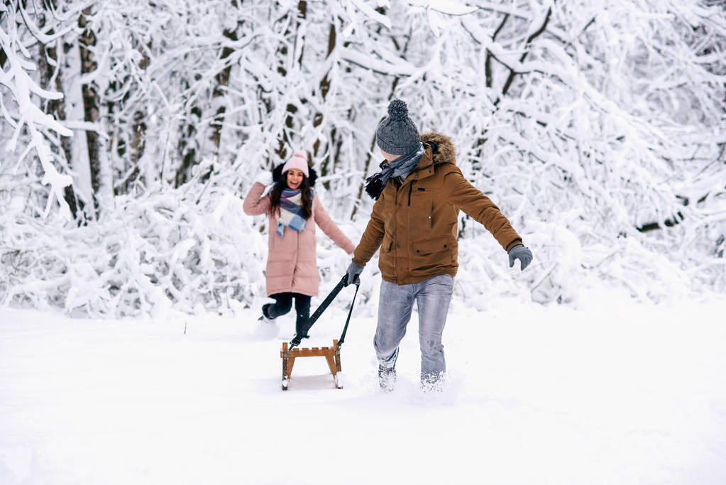 Una hermosa mujer embarazada con ropa de abrigo lanza bolas de nieve a su marido y él con trineos en las manos, escapa de ella. Diversión en el parque de invierno
. - Foto, imagen