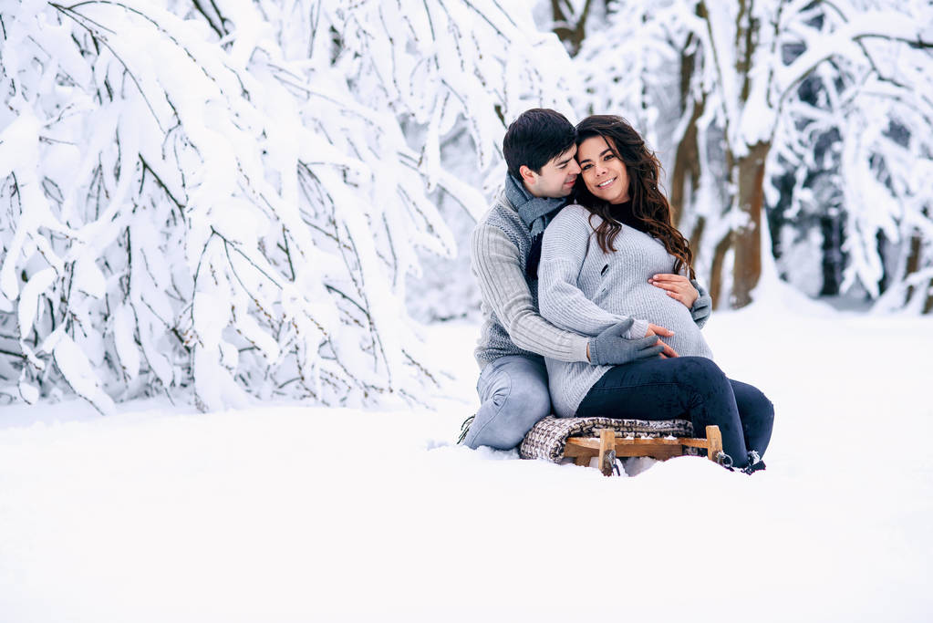 Красивая беременная женщина с мужем, сидящим на санях в снежном зимнем парке
. - Фото, изображение