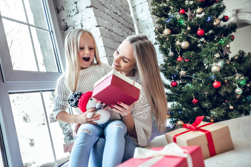 Vrolijke happy vrouw met dochter zittend op de vloer in de woonkamer in de buurt van de kerstboom. New Year's interieur - Foto, afbeelding