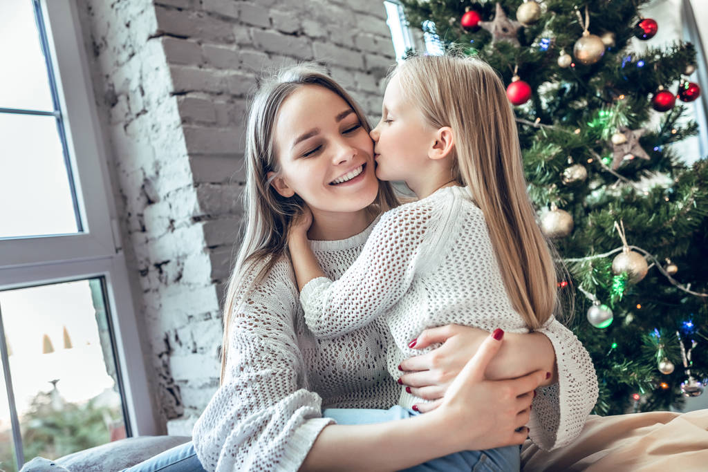 kleines Mädchen küsst seine Mutter auf die Wange auf dem Hintergrund des Weihnachtsbaums. Silvester gemeinsam zu Hause verbringen, glückliches Familienkonzept - Foto, Bild