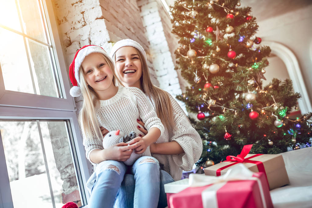 Όμορφη μητέρα αγκαλιάζει κορίτσι Σάντα παιδί και χαμόγελο χαρωπά. Όμορφη κόρη και μαμά στο καπέλο santa στο σπίτι. Καλά Χριστούγεννα και καλές γιορτές! - Φωτογραφία, εικόνα