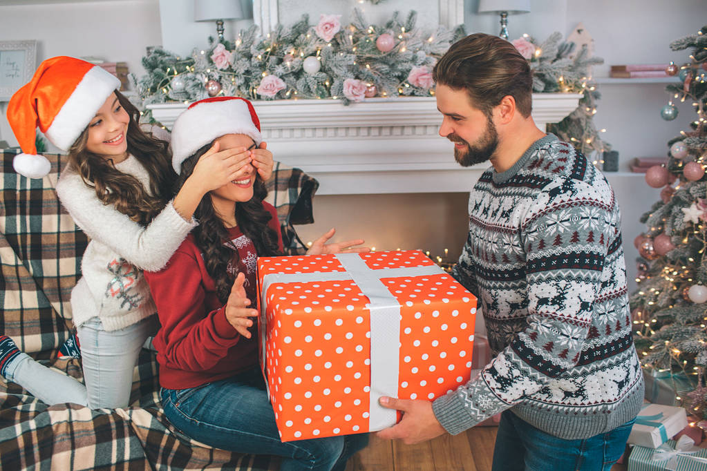Young man and woman holding big box of present. Child kees her mothers eyes closed. Man looks at wife. Family wears festive clothes. - Photo, Image