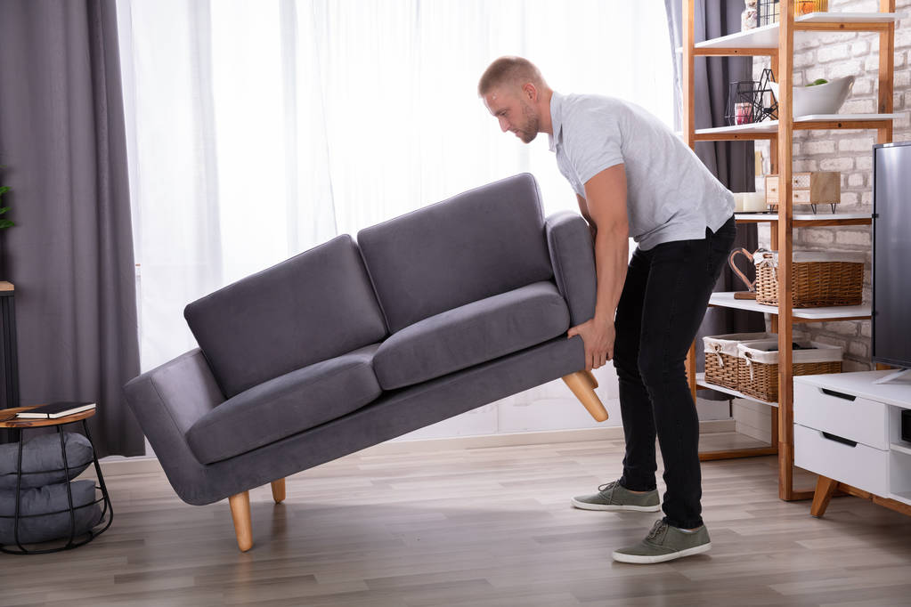 Vista lateral de um jovem homem que levanta o sofá em casa
 - Foto, Imagem