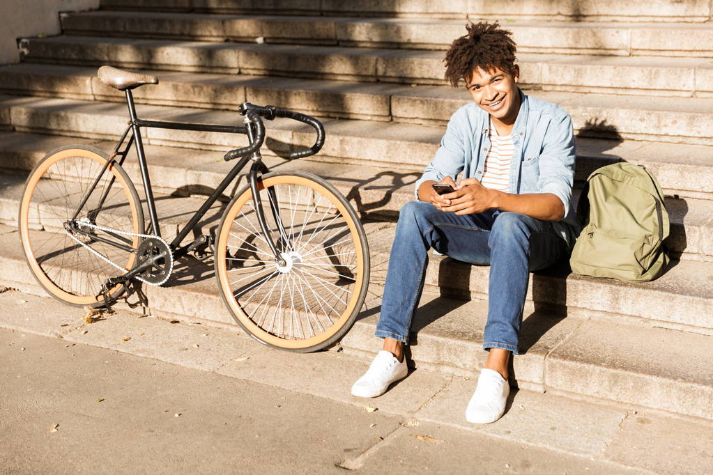 Imagen del joven africano alegre sentado al aire libre con la bicicleta en la calle en las escaleras usando el teléfono
. - Foto, imagen
