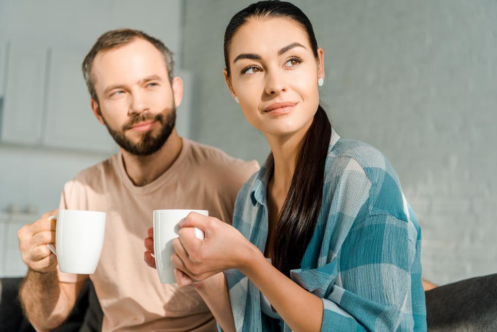 beautiful husband and wife sitting on couch drinking coffee and looking away - Photo, Image