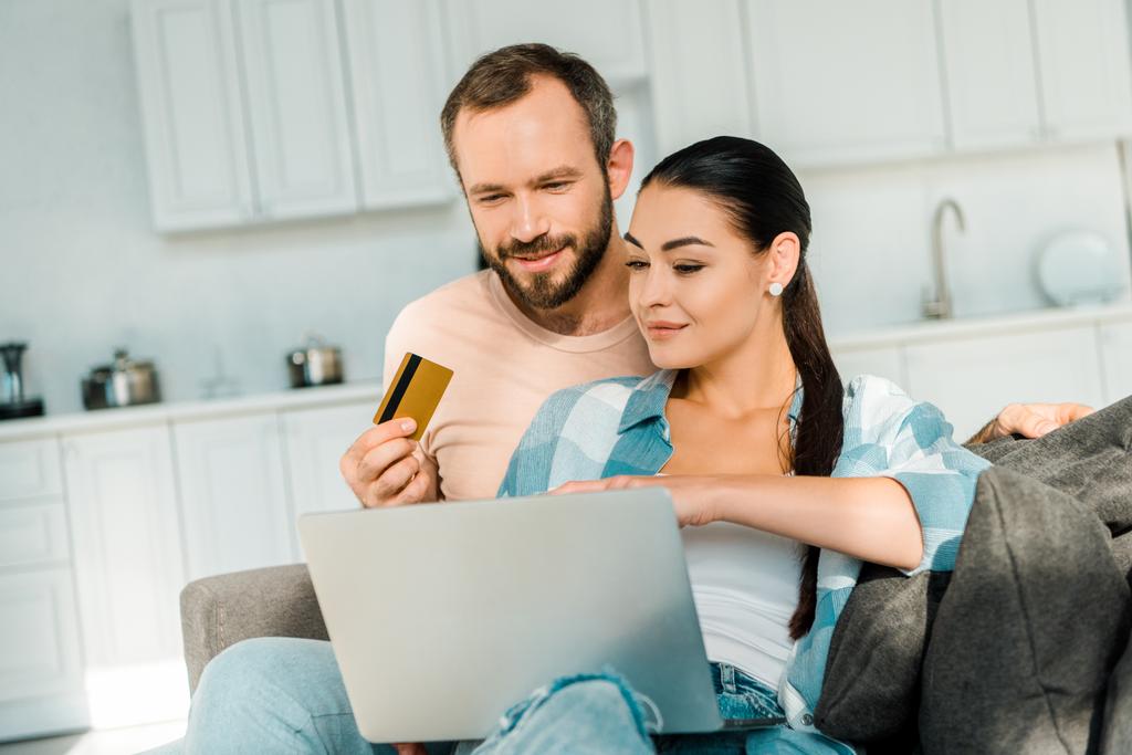όμορφος άντρας κρατάει πιστωτική κάρτα ενώ σύζυγος χρησιμοποιώντας φορητό υπολογιστή και να κάνει online αγορές στο σπίτι - Φωτογραφία, εικόνα