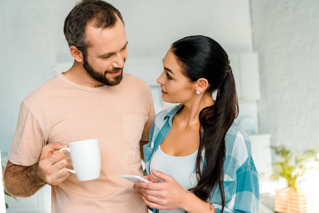 жена с помощью смартфона, улыбаясь муж держа чашку кофе дома
 - Фото, изображение