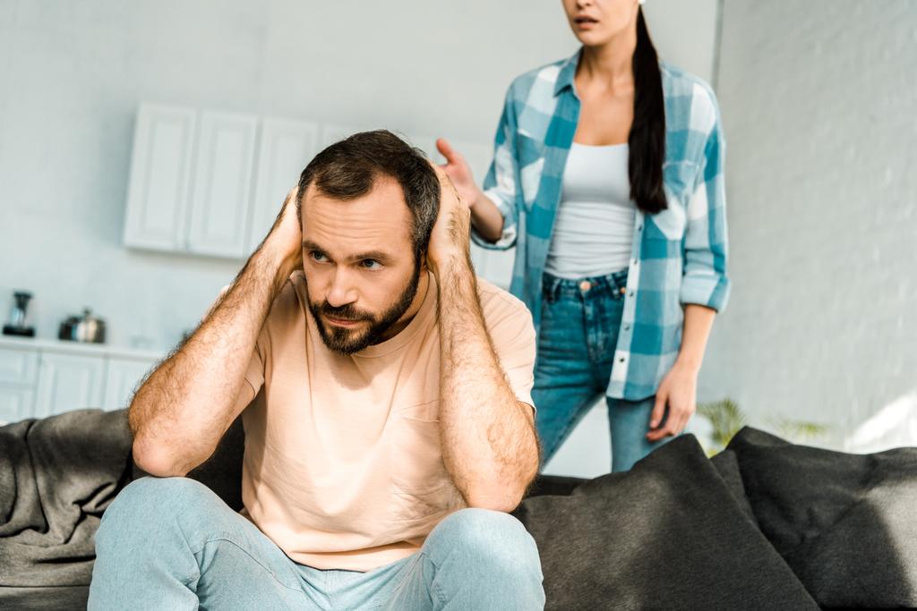 mari stressé au premier plan couvrant les oreilles avec les mains et ayant une dispute avec la femme à la maison
 - Photo, image