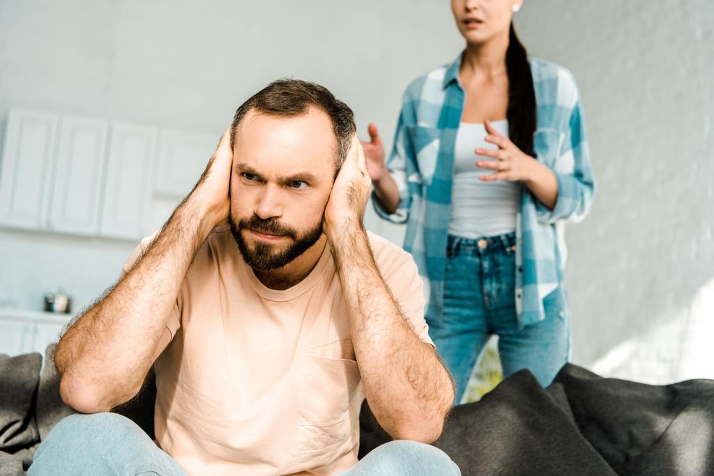 Wütender Ehemann im Vordergrund bedeckt Ohren mit Händen und streitet mit Ehefrau zu Hause - Foto, Bild