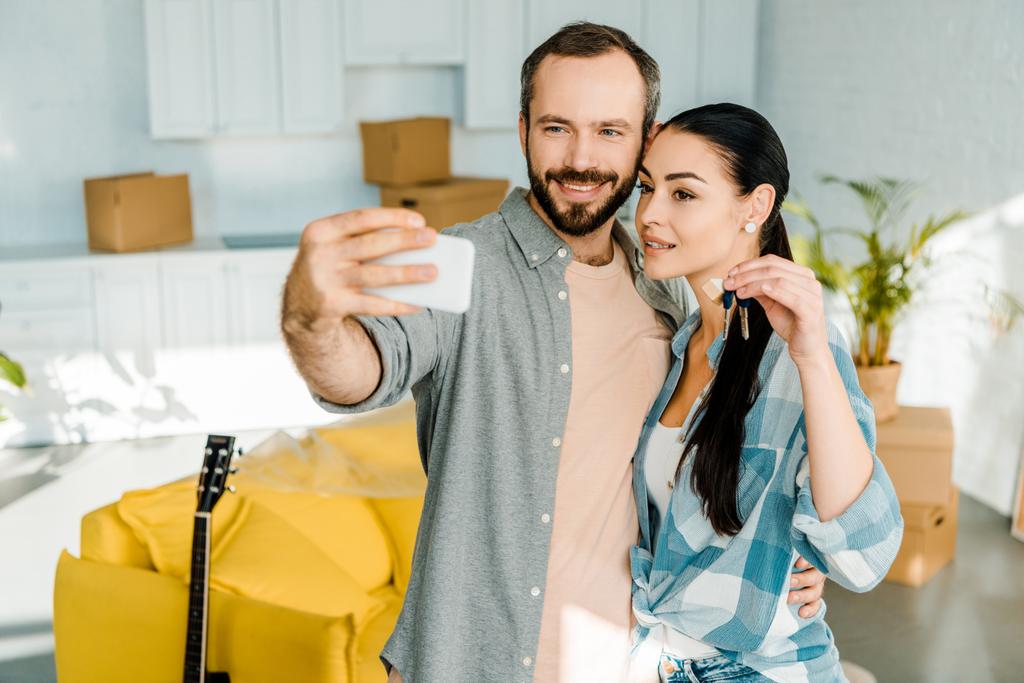 χαμογελώντας σύζυγός λήψη selfie σε smartphone, ενώ σύζυγος κρατώντας τα κλειδιά από το νέο σπίτι - Φωτογραφία, εικόνα
