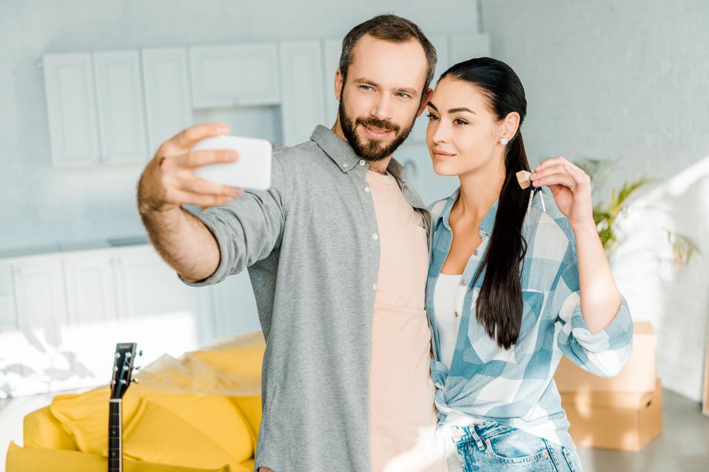 echtgenoot nemen selfie op smartphone terwijl gelukkige vrouw houdt van sleutels uit nieuw huis - Foto, afbeelding