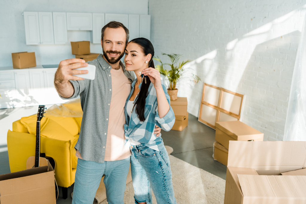 happy moving couple taking selfie on smartphone while wife holding keys from new house - Photo, Image