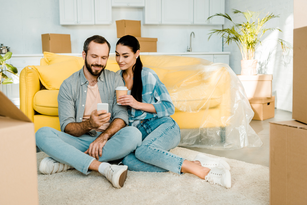 smiling couple drinking coffee, using smartphone and relaxing after packing for new house, moving concept - Photo, Image