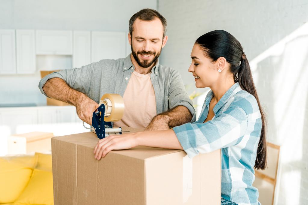 handsome husband and wife packing cardboard box with scotch tape, moving concept - Photo, Image