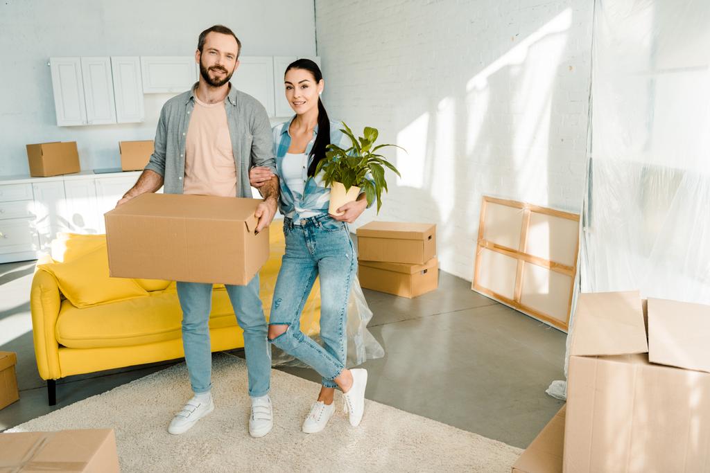 husband carrying cardboard box and wife holding green plant while packing for into new house, moving concept - Photo, Image