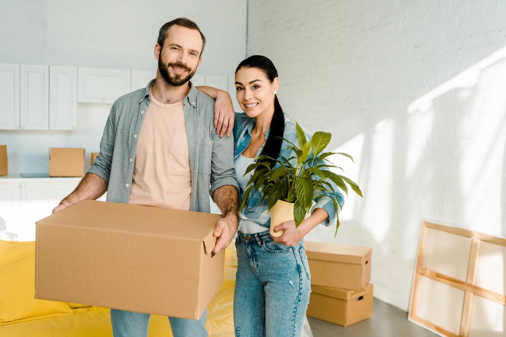 echtgenoot van dragende kartonnen doos en vrouw houden groene plant terwijl verpakking voor nieuw huis, bewegende concept - Foto, afbeelding