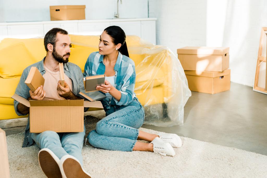 serious couple putting books in cardboard box while packing for new house, moving concept - Photo, Image