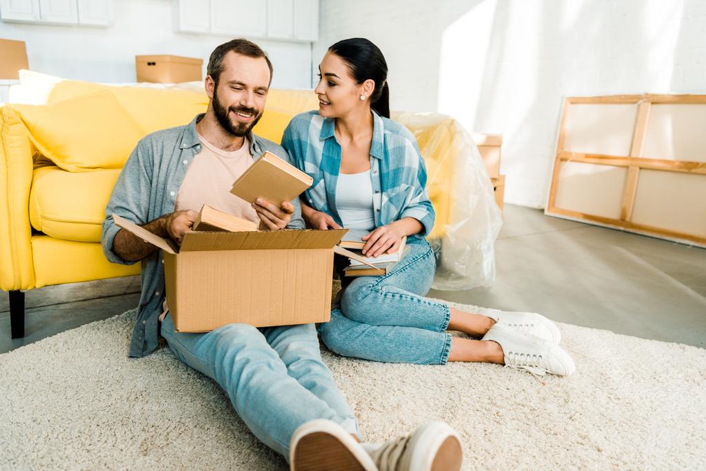 happy couple putting books in cardboard box while packing for new house, moving concept - Photo, Image