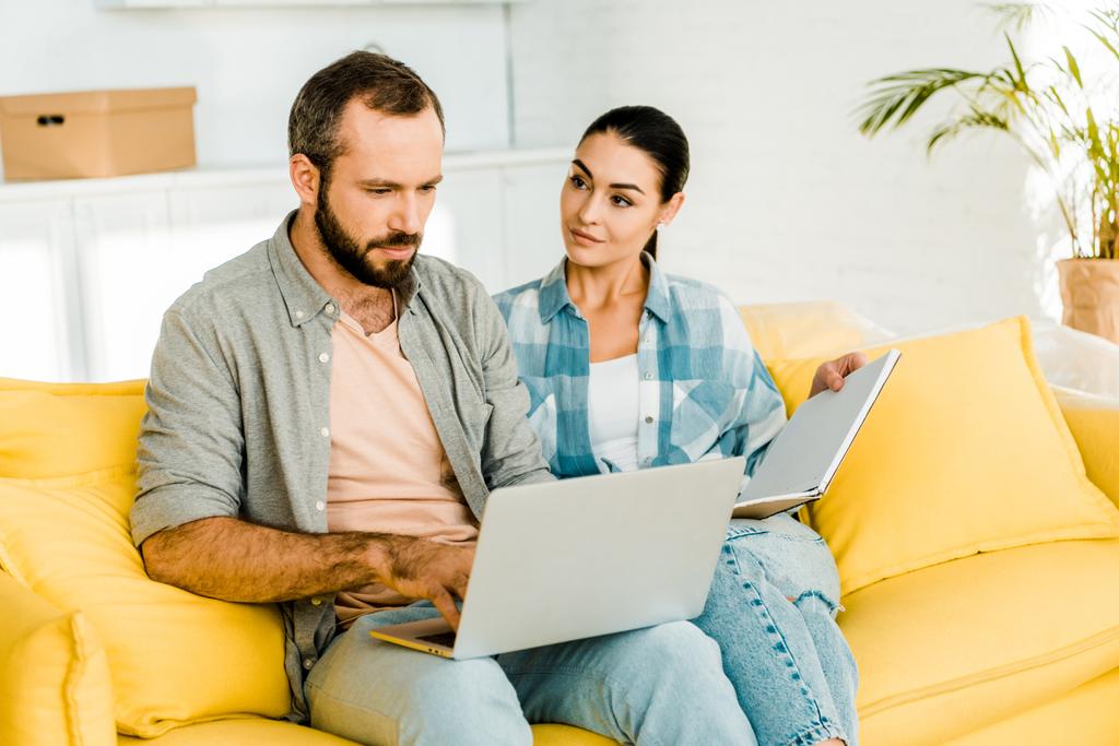 jóképű férje használ laptop, miközben felesége gazdaság notebook, és ül a kanapén - Fotó, kép