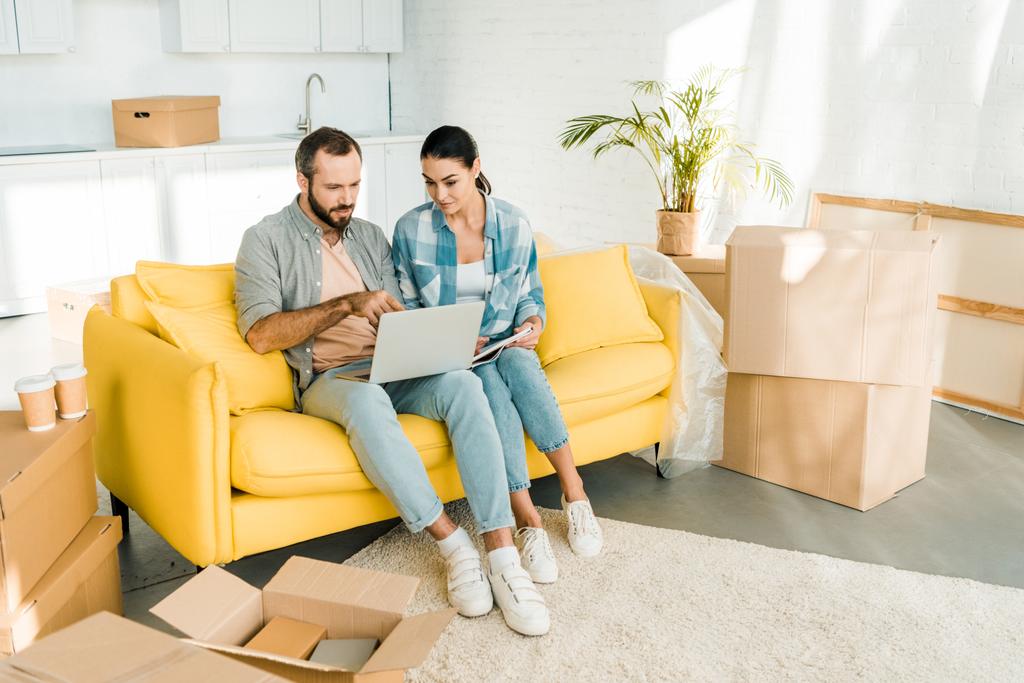 fokussiertes Paar sitzt auf der Couch, nutzt Laptop und plant Umzug in neues Haus, Umzugskonzept - Foto, Bild