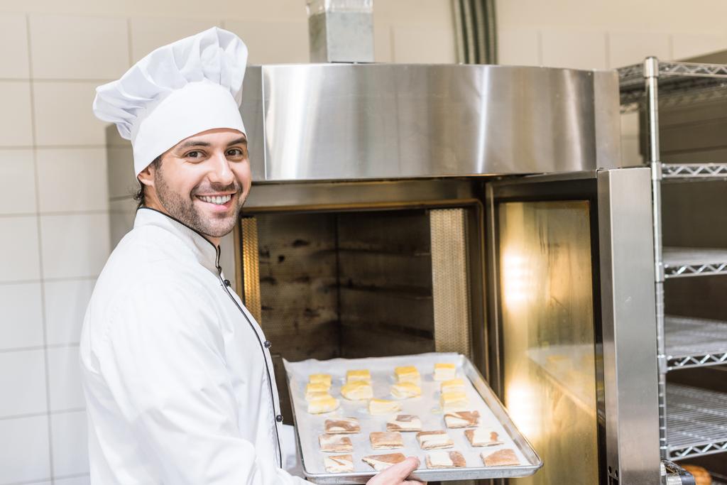 padeiro sorridente em uniforme de chefs brancos colocando bandeja de cozimento com massa não cozida no forno
 - Foto, Imagem