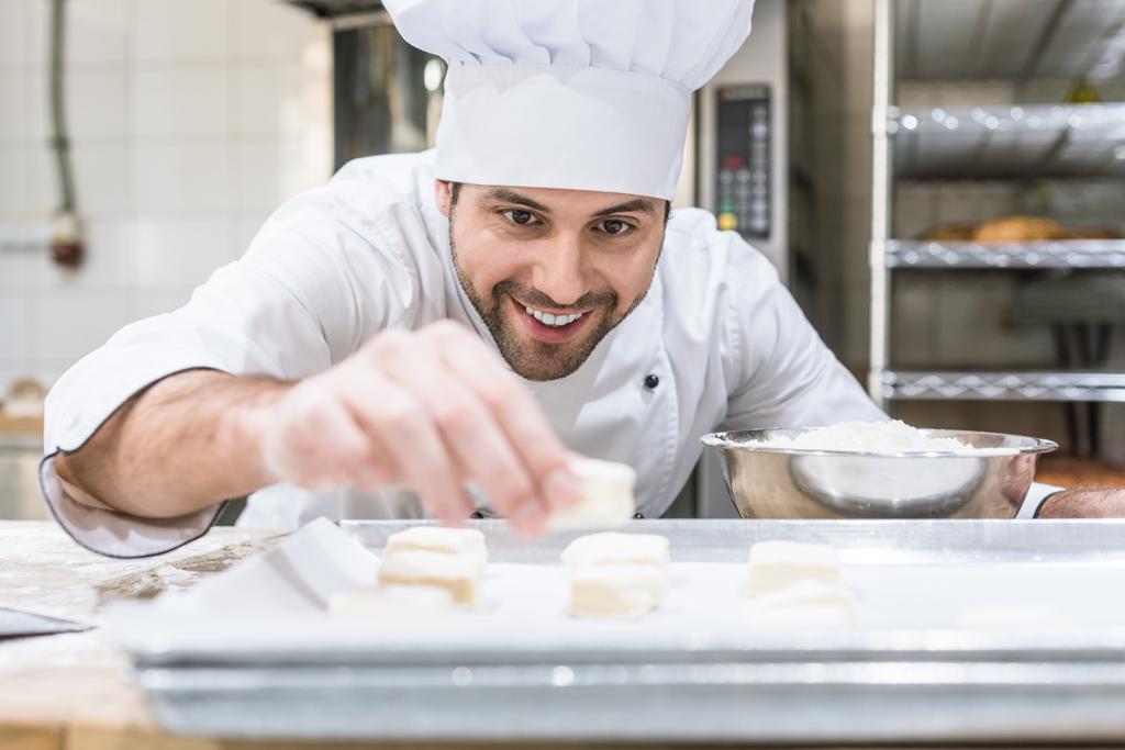Handsome baker smiling and laying uncooked dough on tray - Photo, Image